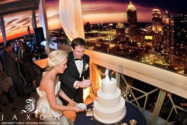 Molly and Dave cuts cake on the roof top at the Peachtree club against a backdrop of the midtown atlanta skyline with a dramatic sunset