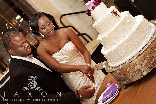 Couple cuts elegant four tiered round wedding cake topped with purple orchids and monogramed in plum and gold