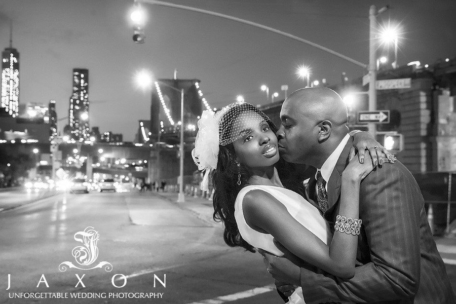 In this night time b&w photograph the groom kisses his bride, the Brooklyn Bridge and mManhattan skyline could be see in the distance | Riviera Brooklyn