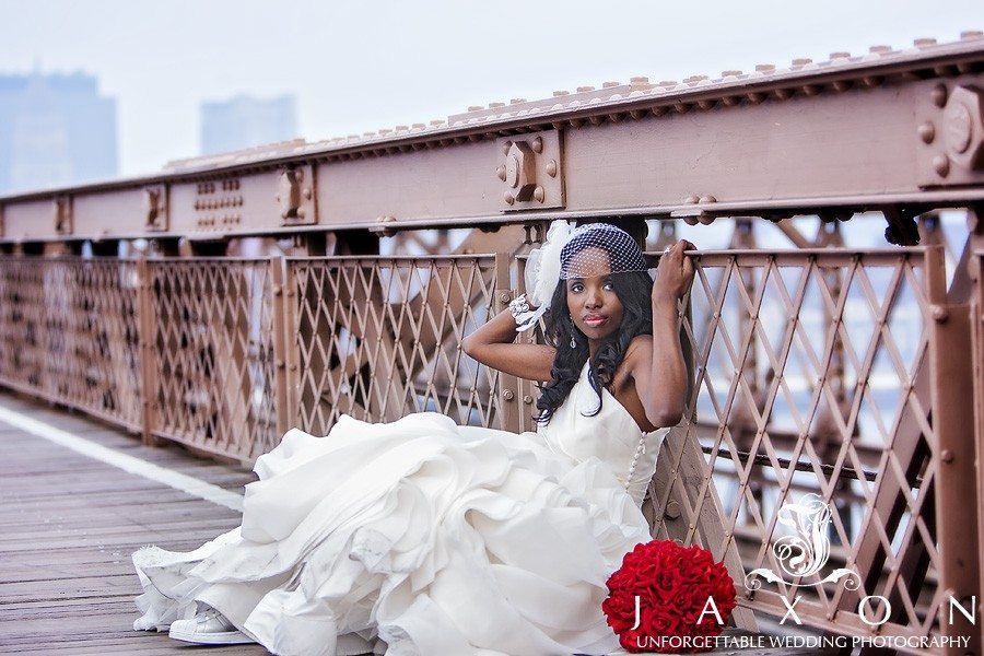 Bride sits on the wooden planks with her back against the railing on the Brooklyn Bridge, her white dress and red rose bouquet contrasts beautifully against the rust colored Brooklyn Bridge | Riviera Wedding Brooklyn, NY