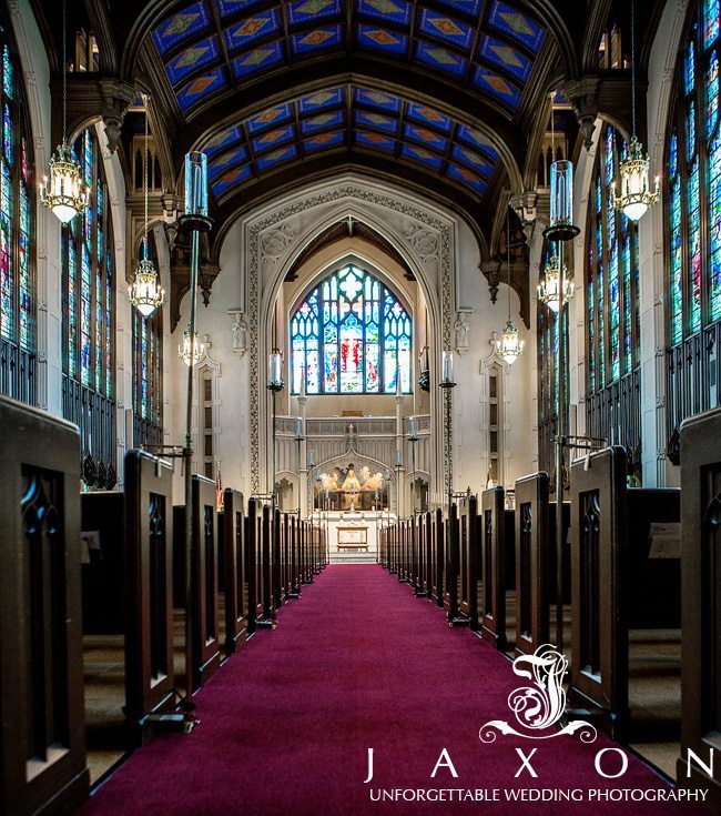 Inside the cathedral with the aisle paved in maroon carpet, side tall light blue stained glass windows and an arched roof with darker blue stained glass windows