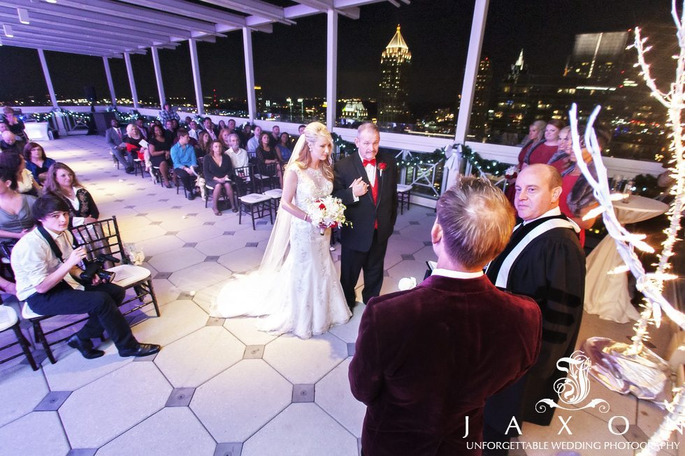 Father escorts bride into her wedding ceremony on the rooftop of the Peachtree club, high rise buildings glimmers in the night sky