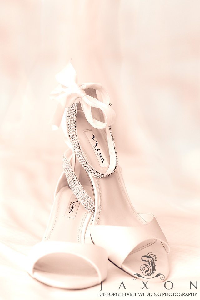 Sepia photo of white Nina NY Crystal Embellished Ankle Strap Sandal with bow, on bed at the Loews Hotel Atlanta 