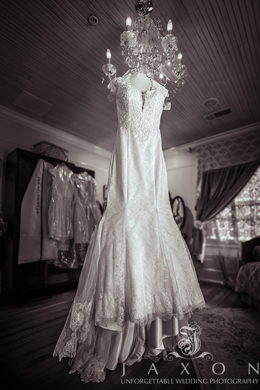 Galina Signature trumpet wedding gown with a plunging V-neckline hanging from a chandelier in Carl House bridal suite, sepia-toned with bridesmaids' dresses in the background.