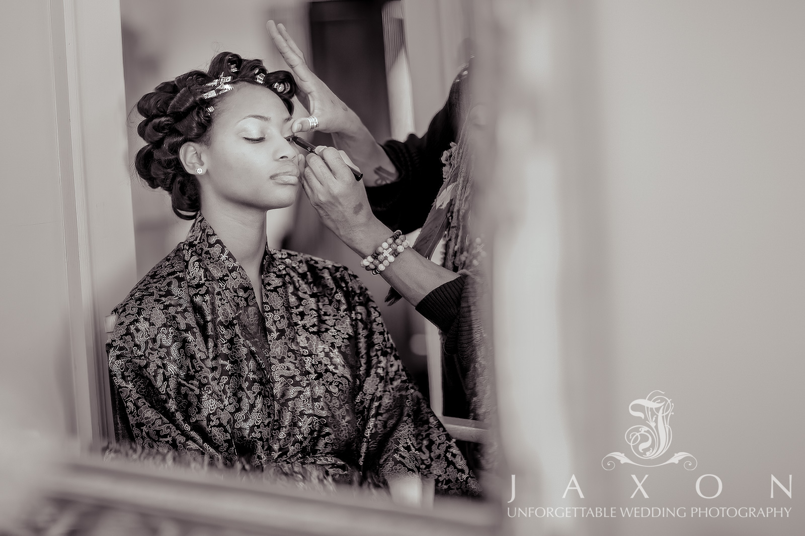 Sepia-toned photograph of a makeup artist applying eyeliner to a bride in a silk robe, with pinned-up hair, at Carl House, a wedding venue in Atlanta.