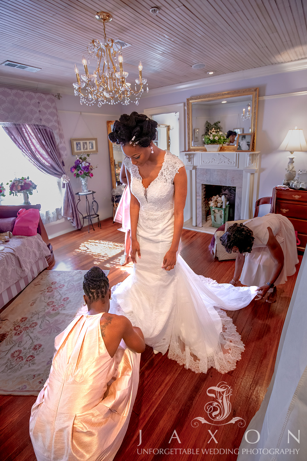 Bride being assisted by two attendants in a luxurious Carl House bridal suite, wearing a Galina Signature tulle-over-lace trumpet gown with a plunging V-neckline and cap sleeves.