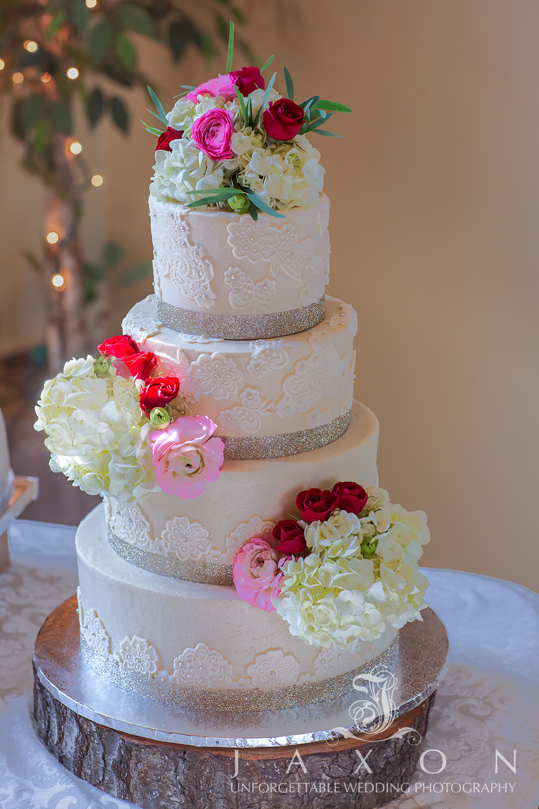 A beautifully crafted four-tiered round wedding cake with white lace brushed embroidery and silver bands at the base of each tier, adorned with clusters of white anemones, red roses, white French tulips, hydrangeas, and ranunculus blooms, displayed in the Carl House ballroom in Auburn, Georgia.