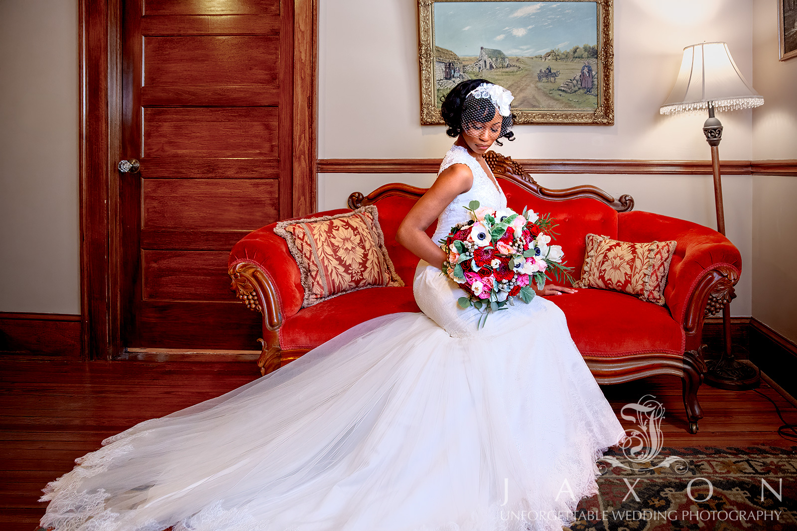 Bride in Galina Signature tulle-over-lace trumpet gown with a flower-topped birdcage veil, seated on a red velvet loveseat with a bouquet, in the Carl House Mansion bridal suite.