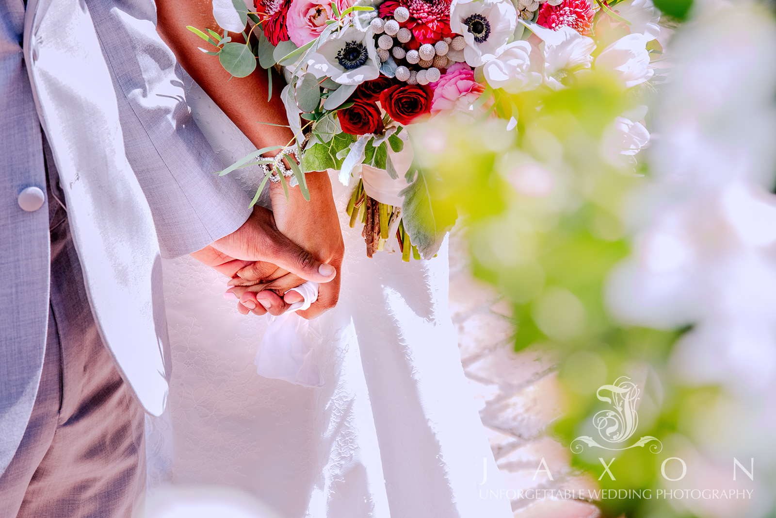 Close-up of couple holding hands through foliage during wedding ceremony, highlighting bride's white dress and groom's light gray suit.