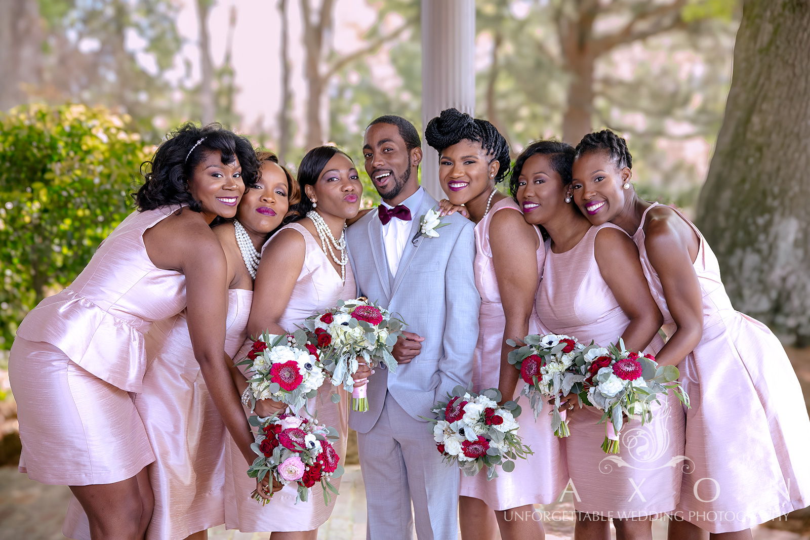 Groom in gray tux and plum bowtie with smiling bridesmaids in blush pink dresses holding colorful bouquets.