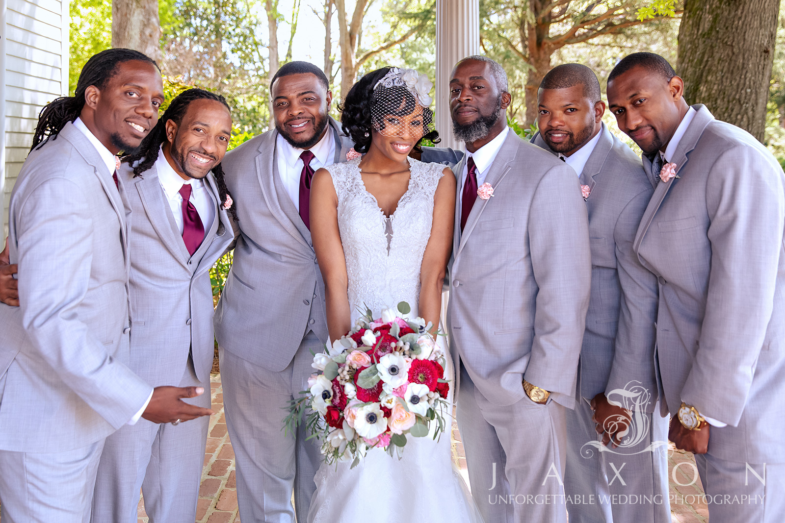 Bride with groomsmen in gray tuxedos and burgundy ties, holding a white and red floral bouquet.